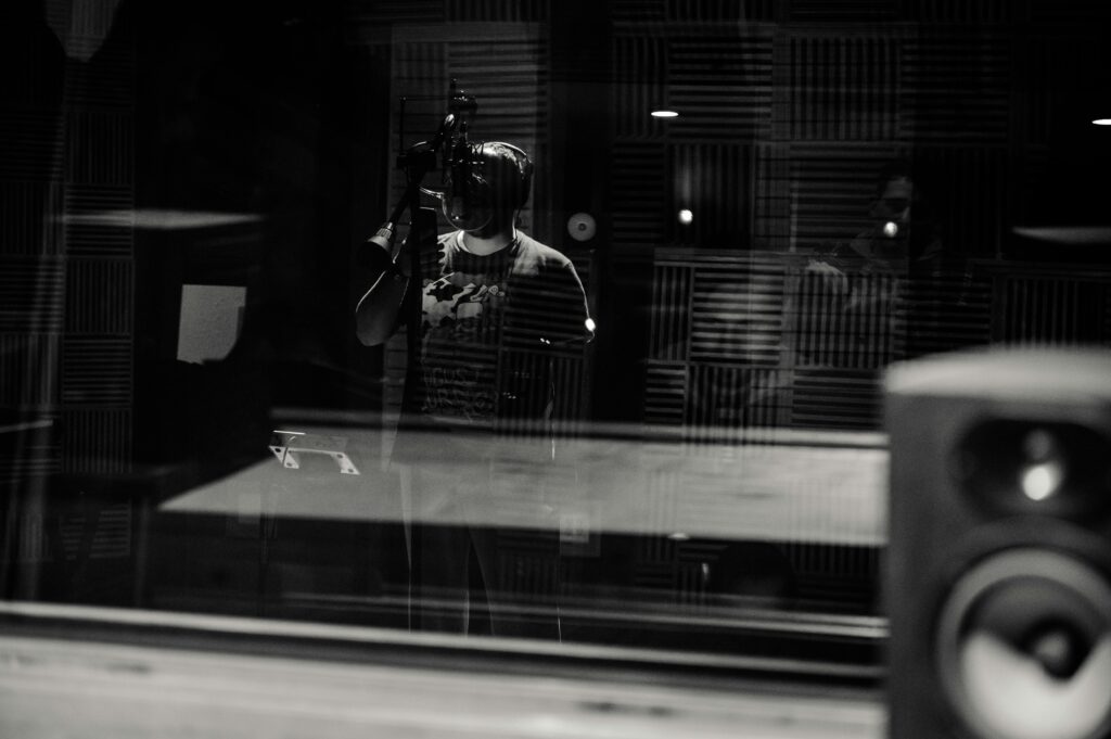 pexels-photo-4143428-4143428 Through glass of black and white anonymous male musician in headphones singing with microphone while recording song in modern studio